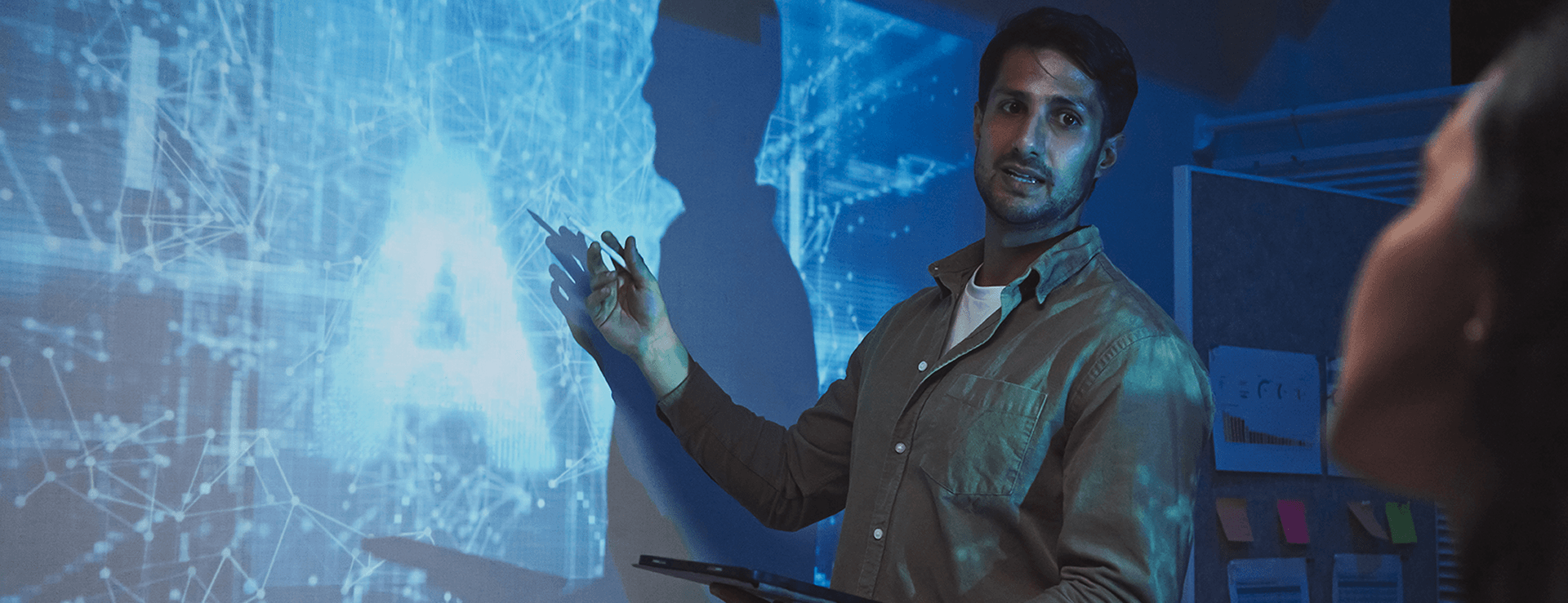 A man in a shirt points with a pen at a wall projection showing the word "AI" (artificial intelligence) and a blue, networked pattern while giving a presentation.