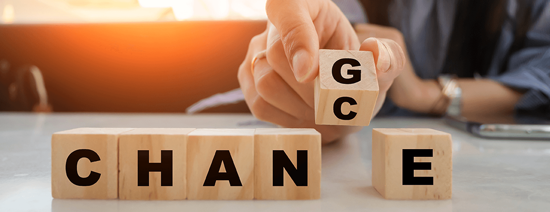 Close-up of a hand inserting a wooden cube with the letters G and C into the gap of the word CHAN E on a table. The row of cubes spells out the word CHANGE, and the inserted cube can form the word CHANCE, symbolizing that every change is an opportunity.