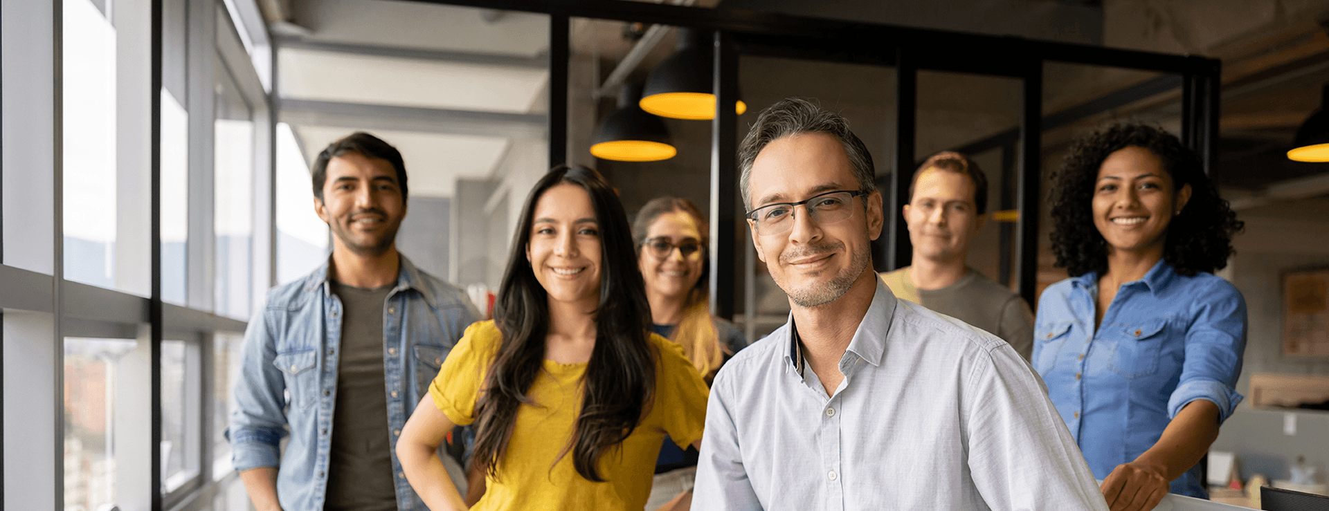 A diverse group of six smiling colleagues pose confidently in a modern office with large windows in the background.