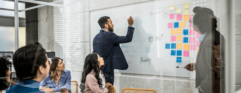 A businessman in a suit writes with a marker on a large, transparent whiteboard wall in a modern office while a group of colleagues look on during a meeting.