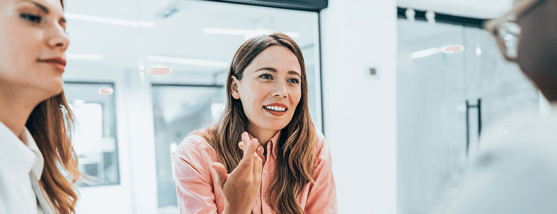 Smiling young businesswoman in a pink shirt gestures with her hands while talking to colleagues in a modern office environment.
