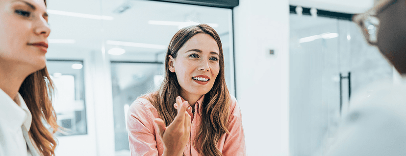 Smiling young businesswoman in a pink shirt gestures with her hands while talking to colleagues in a modern office environment.