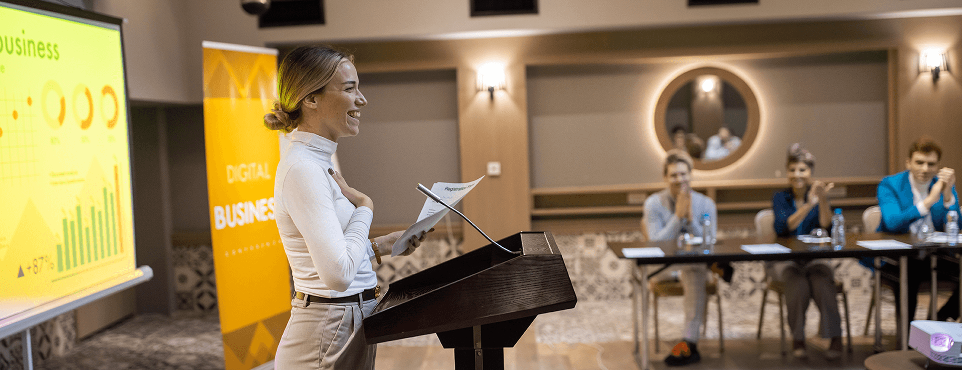 Blonde, smiling speaker in a white turtleneck sweater gives a presentation while the applauding audience sits at a table.