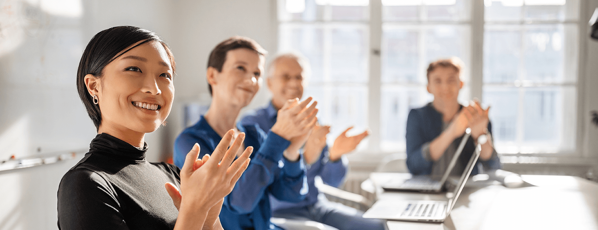 Smiling woman with dark hair in a black turtleneck sweater gossiping with three blurry, applauding colleagues at a conference table with laptops near a bright window.