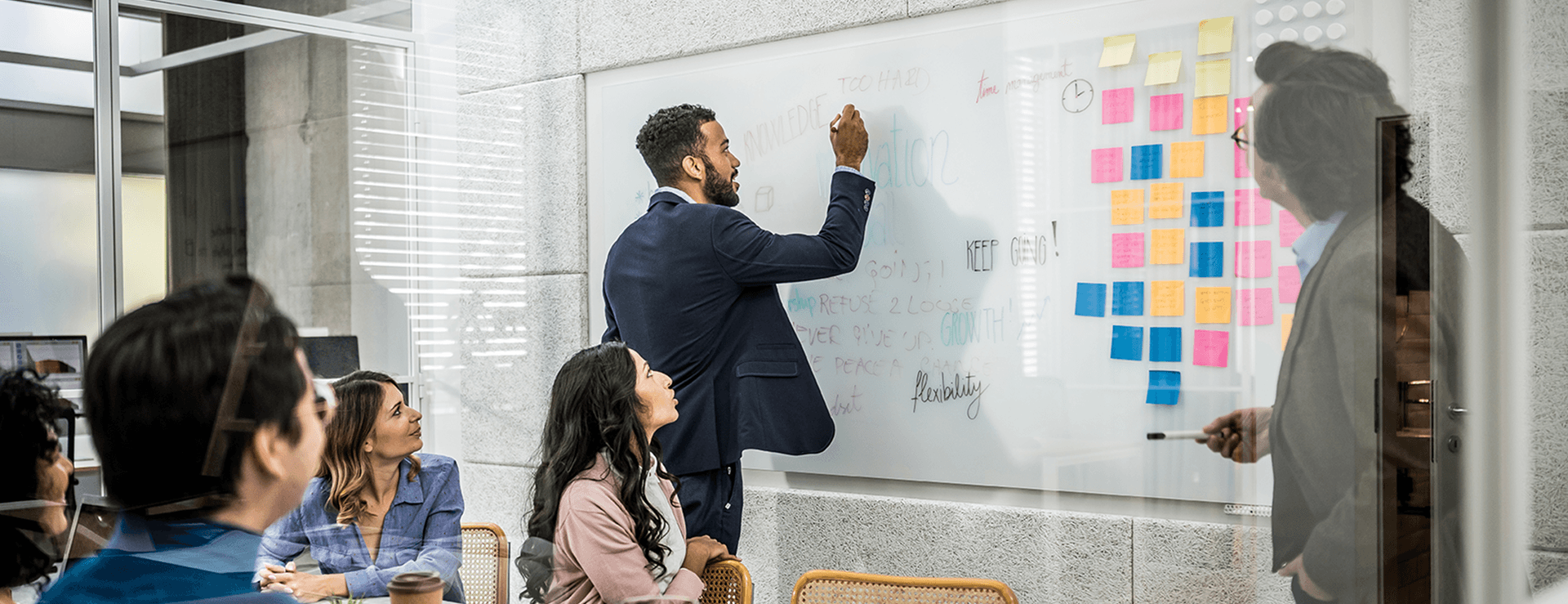 A businessman in a suit writes with a marker on a large, transparent whiteboard wall in a modern office while a group of colleagues look on during a meeting.