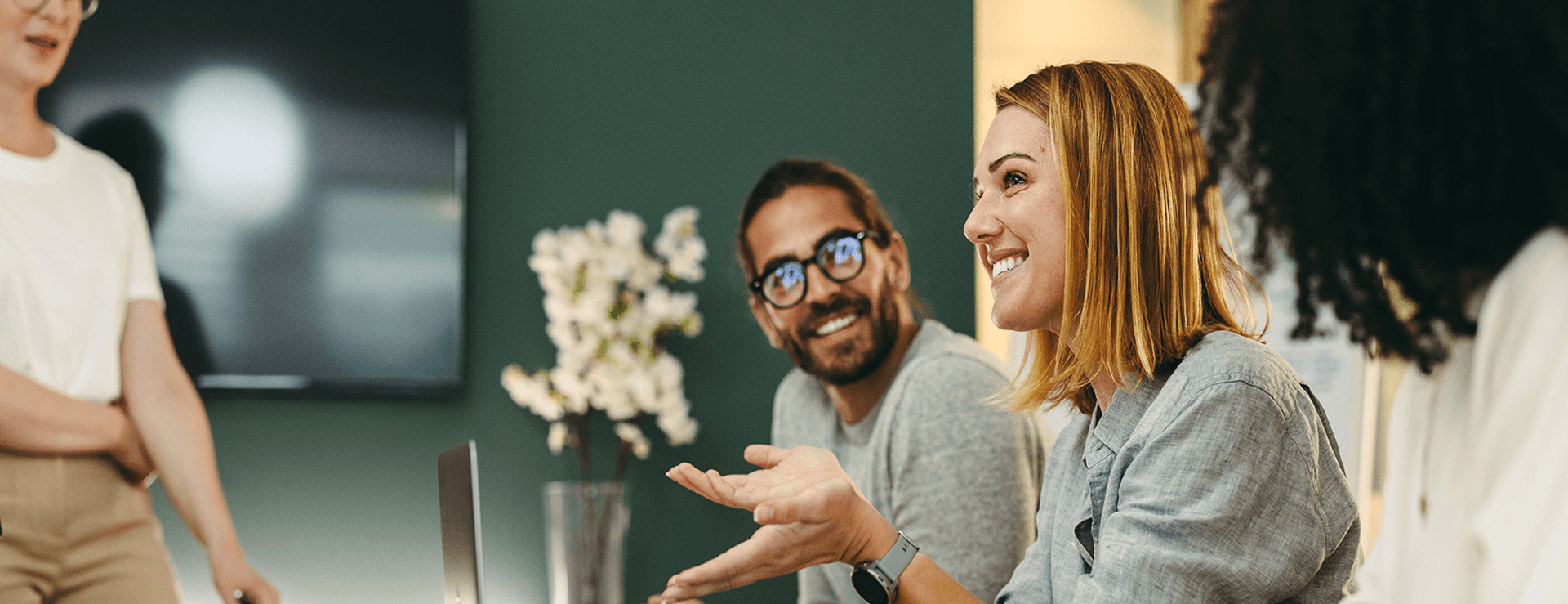Smiling businesswoman in gray shirt speaks and gesticulates in a meeting; bearded colleague with glasses listens attentively.