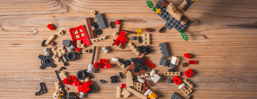 Verstreute, bunte Lego-Bausteine, darunter rote, beige, graue und schwarze Teile, sowie eine unfertige graue Konstruktion auf einem Holztisch.